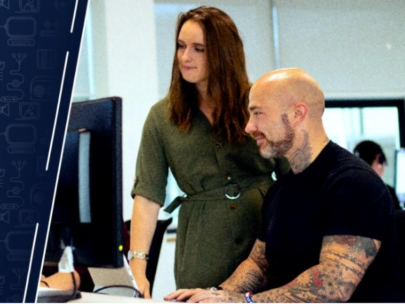 A photo of A man and a woman looking at a monitor together. The woman is wearing a green dress and the man is wearing a black tshirt and has arm tattoos