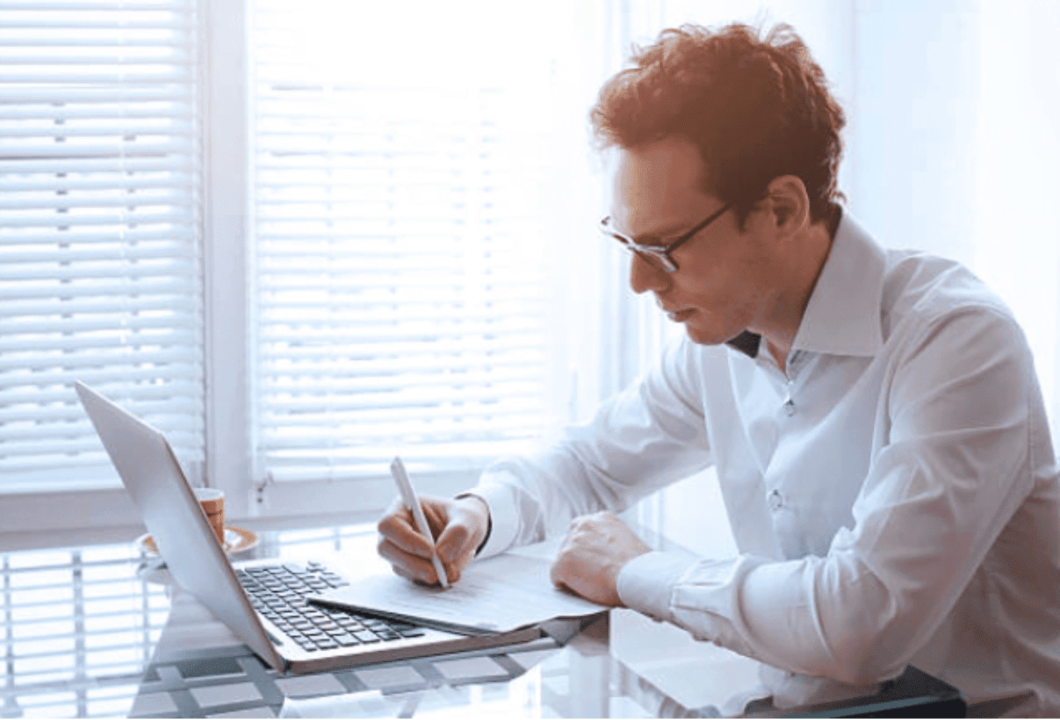 A man writing on a pad of paper, whilst looking at his laptop