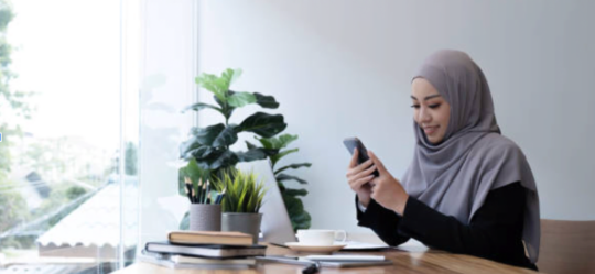 A lady wearing a hijab looking at her mobile phone whilst sat at a desk