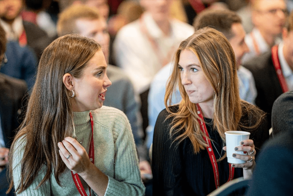 Two female colleagues sitting in the audience at an event. they are chatting and drinking coffee.