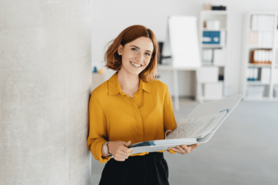 Photo of a young apprentice in an office. She is wearing a yellow blouse and holding an open ring-binder