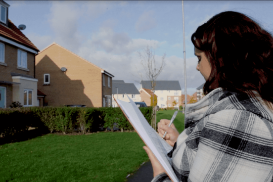 A site inspector completing a form outside a house, she has dark hair and is wearing a plaid coat.