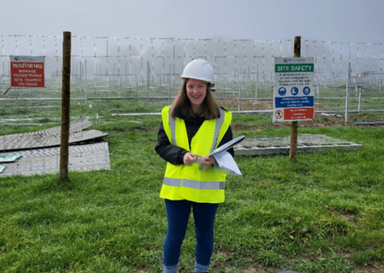 Decorative image: A lady called Jess, at a construction site. She is wearing a hi-vis jacket and a hardhat.