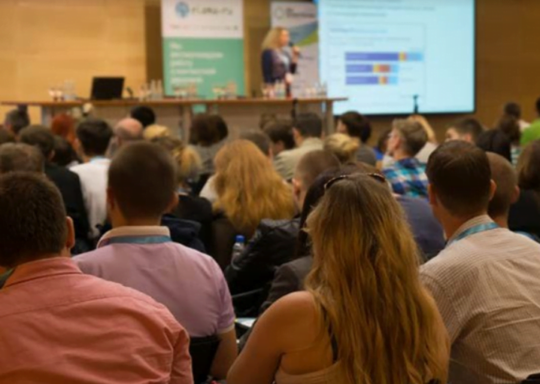 Decorative image: a photo of a lecture hall full of people, all looking at a presentation screen