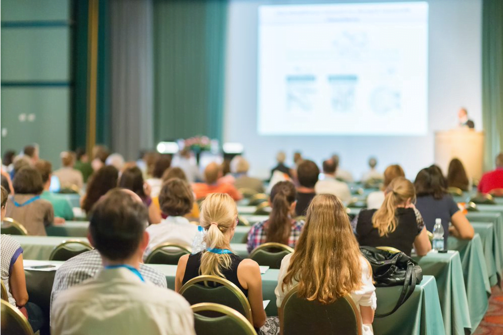 Decorative image: rows of people at a conference, all looking away from the camera, towards a screen with graphs on