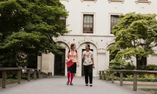 Two female colleagues strolling away from a building, down a tree-lined path
