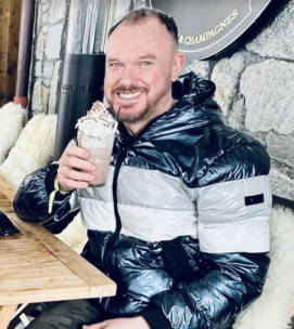 Lyle Pewsey sits at a table with a milkshake in one hand smiling at the camera