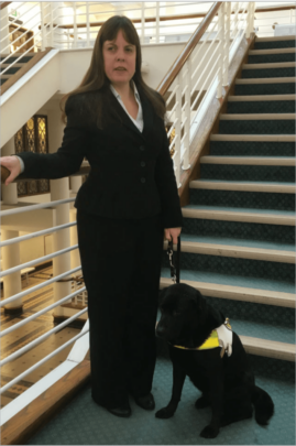 Lisa Boocock and her guide dog Bess. Lisa is standing by a flight of stairs and is dressed in a smart suit, Bess in a black labrador.