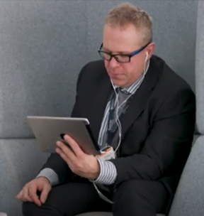 Aidan McGuffie. He is sitting down and focused on a tablet device, listening through headphones. He is wearing a dark suit and pale blue shirt.