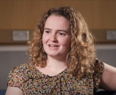Danielle Davies. She has shoulder length curly hair and is wearing a floral top.