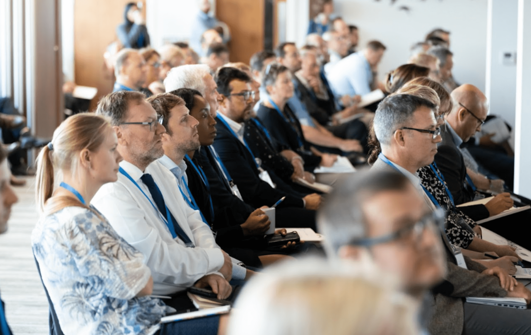 An audience at an event. They are professionally dressed, wuth matching blue lanyards, and are sitting listening to a presentation.