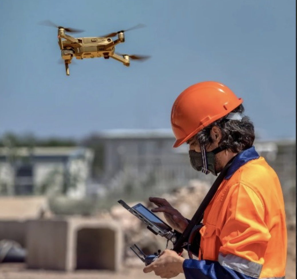 A person in a safety helmet, using a tablet device to fly a drone which hovers in the background