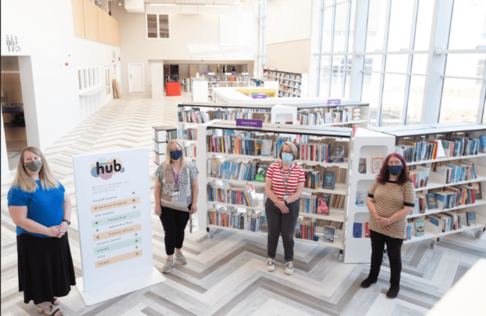 Four women standing a brightly lit library. They are wearing facemasks, and are socially-distanced, indicating that this was during the pandemic.