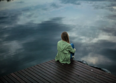 A woman sitting on a wooden dock next to a lake, in winter clothing.