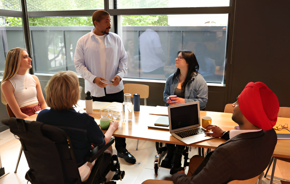 A group of colleagues in a meeting. One man is man standing to give a presentation, two female wheelchair users, and a man wearing a red turban are listening intently.