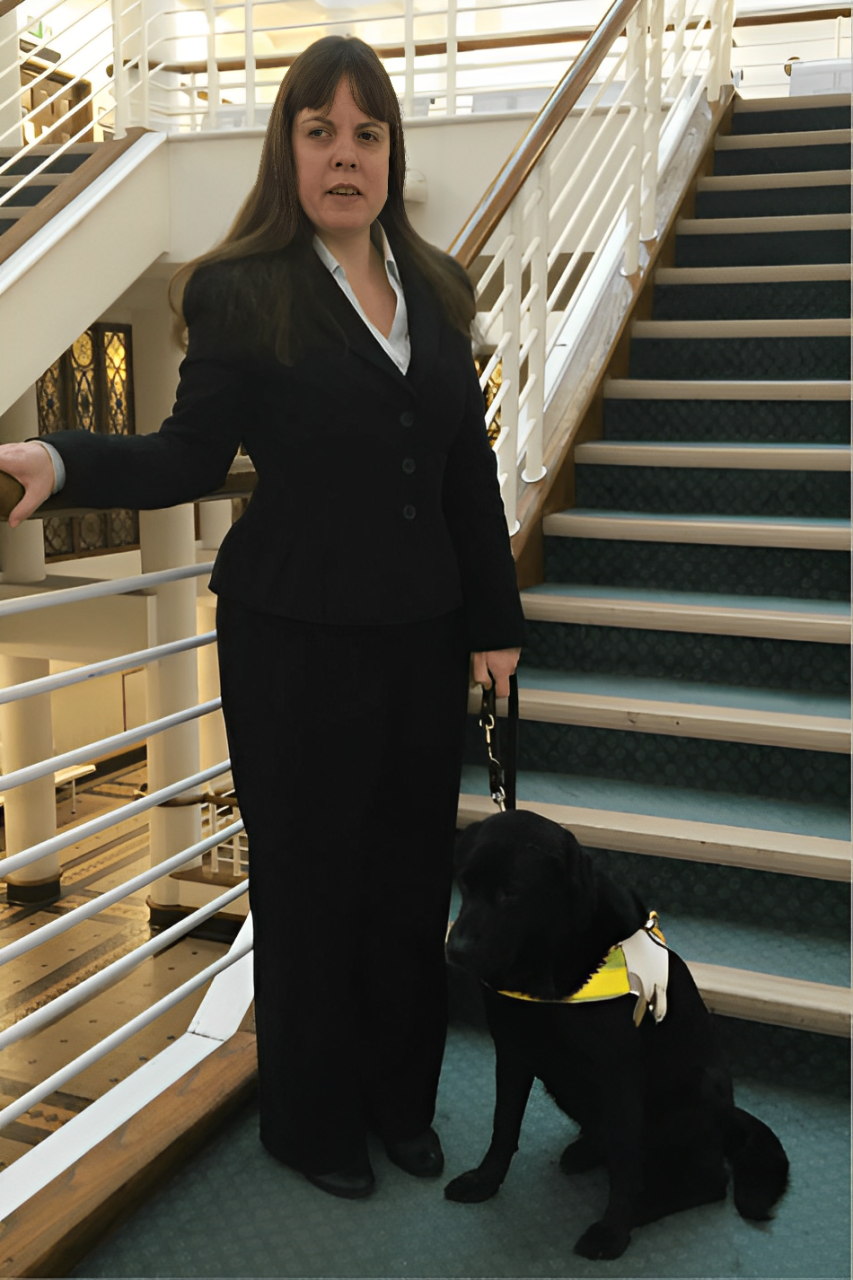 Lisa Boocock and her guide dog Bess. Lisa is standing by a flight of stairs and is dressed in a smart suit, Bess in a black labrador.