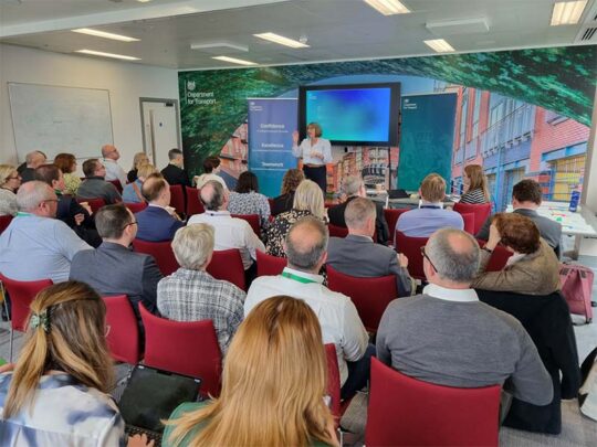 A presenter speaking to a group of people about confidence, excellence and teamwork in the context of leadership.