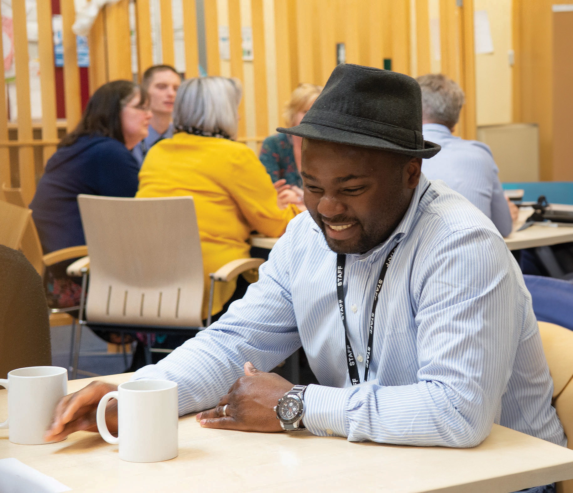 Man with a trilby hat sitting at a table smiling with someone that is out of shot. Behind him are a group of people sitting around another table appearing to be in discussion with each other.