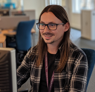 Photo of Steven Banks, who is a Level 6 Digital And Technology Solutions Apprentice at the Ministry of Defence. He has long brown hair and is wearing dark-rimmed glasses and a plaid shirt