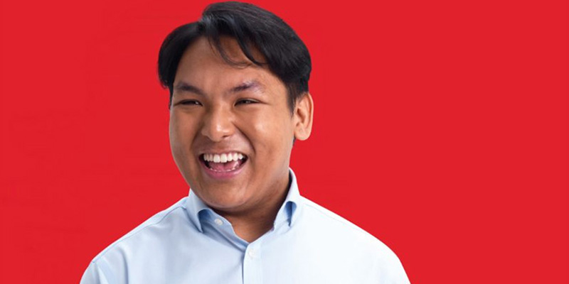 A young man smiling. He is wearing a button-down blue shirt and is in front of a bright red background.