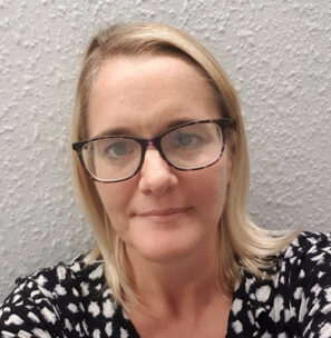 Photo of Suzanna Hanna an Operational Lead in DWP. She has shoulder-length blonde hair, black-rimmed glasses and is wearing a patterned blouse