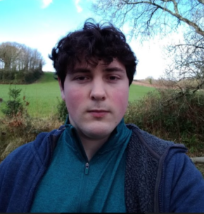 Photo of Thomas Dufall. He has short dark hair and is pictured outside, wearing a jumper and coat in blue tones