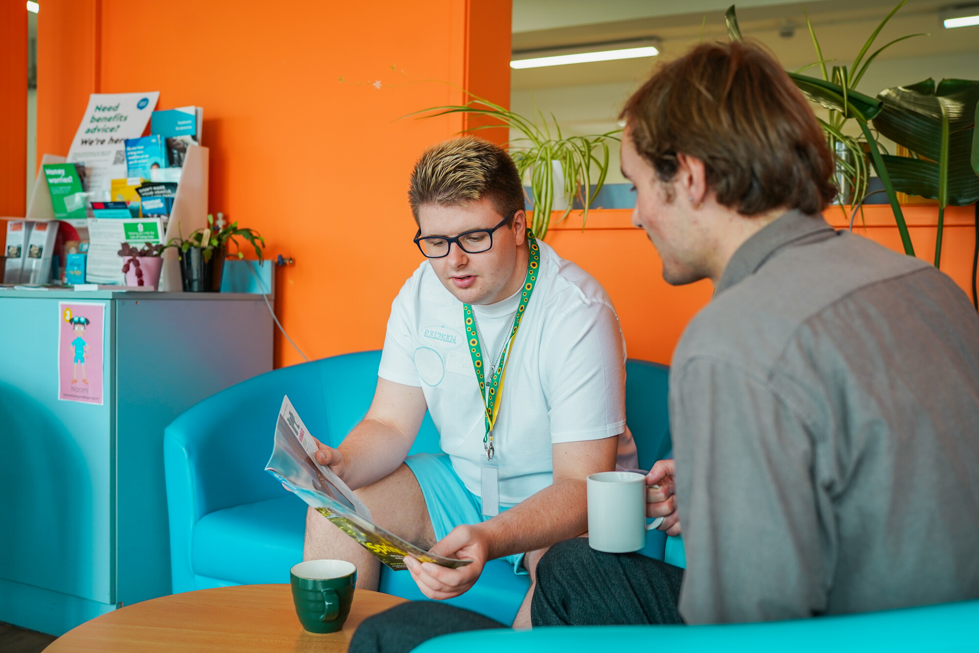 Two men with less visible disabilities are in a break-out room, reading a magazine and drinking tea together. One man is wearing a sunflower lanyard.