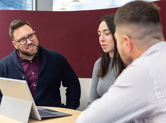 3 colleagues sat round a table with a laptop