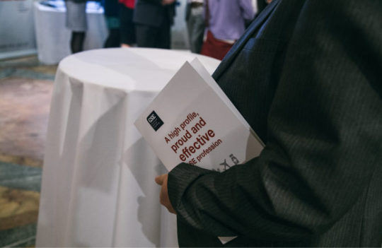 The torso of a person in a suit jacket, holding a pamphlet at an event