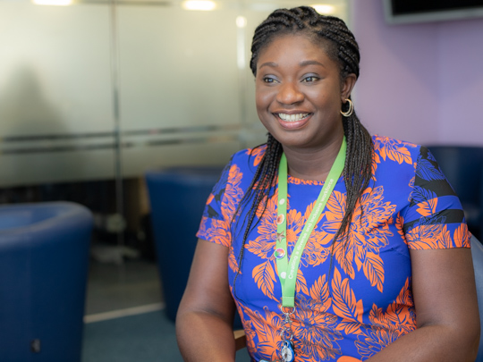 A photo of Uche Williams. She is wearing a brightly pattered top in blue and orange and has dark braided hair. She is smiling.