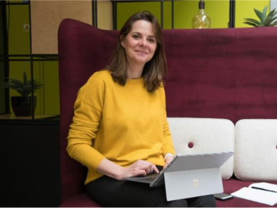 A photo of Jennifer, she is wearing a yellow jumper smiling at the camera while working on her laptop