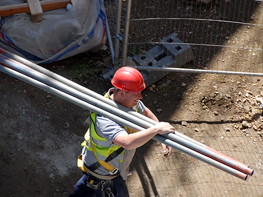 Photo of a builder in a hardhat. He is carrying several long metal pipes over one shoulder.