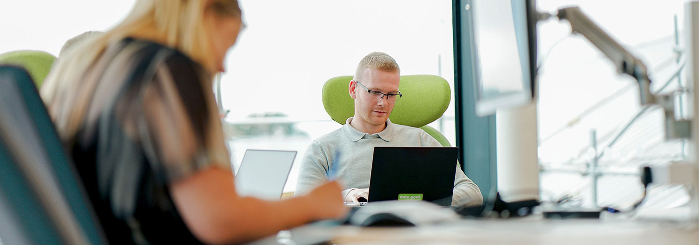 Two colleagues, sitting at a table, working on laptops. The room is open and light.