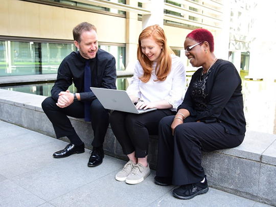 Decorative image: three colleagues sitting outside together, looking at a laptop. They are dressed in smart-casual clothing and smiling
