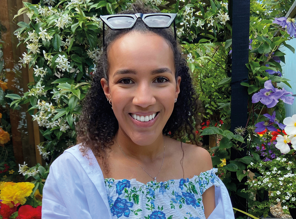 A photo of Hannah. She is wearing a floral top and has her sunglasses perched on her head. She is surrounded by flowers and plants.