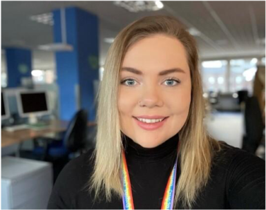 A photo of Holly. She has blonde hair and is wearing a black sweater and a rainbow lanyard.