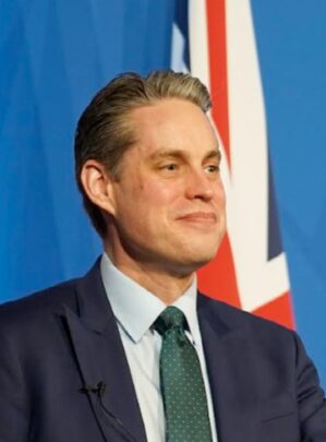 A photo of Simon Baugh. He is wearing a dark suit and standing beside a union flag.