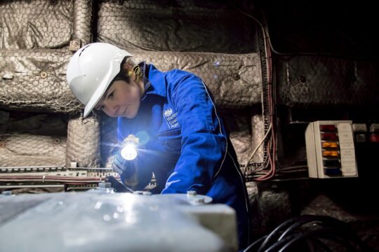 Surveyor, Leanne Grillandini, at work. She is wearing a white hardhat and blue coveralls. She is using a torch to inspect a piece of machinery.