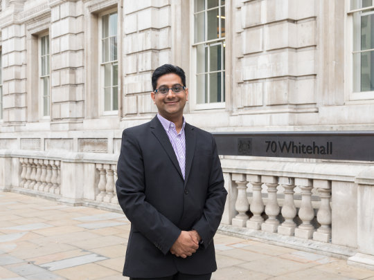 A photo of Nasser Sheikh Outside 70 Whitehall. He is smiling and wearing a dark suit with a lavender shirt.