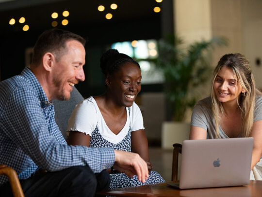 Three diverse colleagues sitting together in a brightly lit modern office lounge, smiling as they collaborate and look at a laptop screen on a low table.