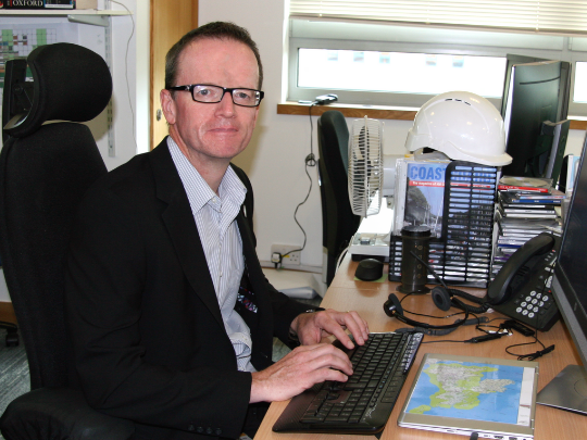 A photo of Rob Townsend. He is sitting at a desk and is dressed in a suit with a white shirt.