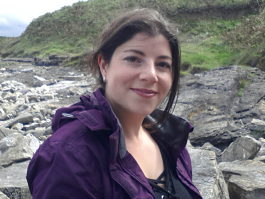 Photo of Ruth Kyriacos, a Dvsa Vehicle Examiner. She is wearing a purple windbreaker and is pictured outside in front of a rocky outcrop