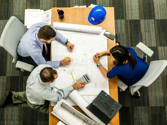 Three HM Treasury team members, working around a table which has a large piece of paper, a hard hat and a laptop on it