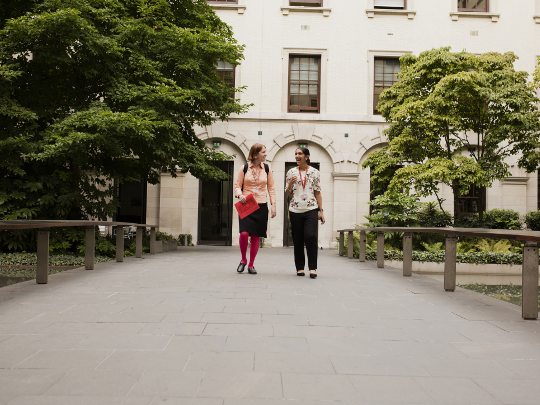 Two female colleagues strolling from a building, down a tree-lined path